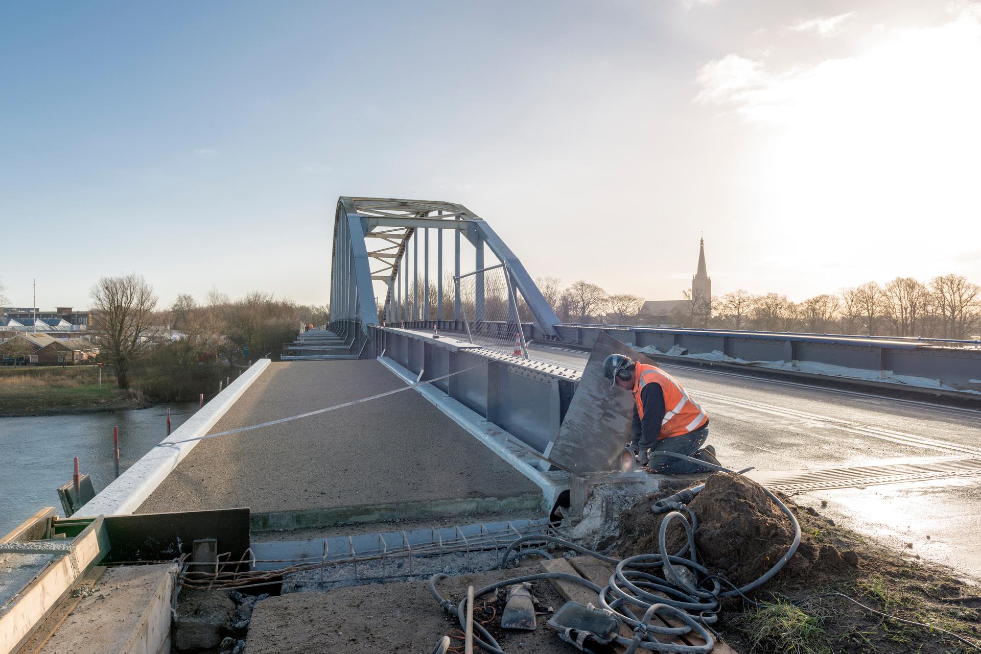 Een bouwvakker met een slijptol op een brug over de rivier. Op de achtergrond is bebouwing.